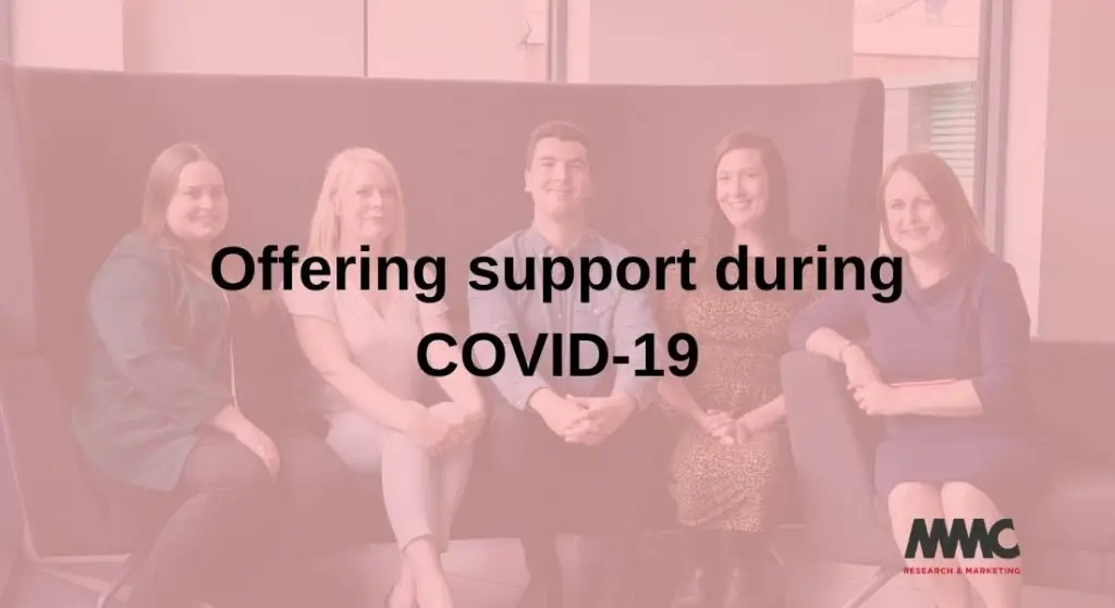 A wide angle photo of 5 team members of MMC sat on a high-back couch wearing business dress in a business environment. The image has a red wash over it and is faded. Large black text is written over the centre of the image "Offering support during COVID-19