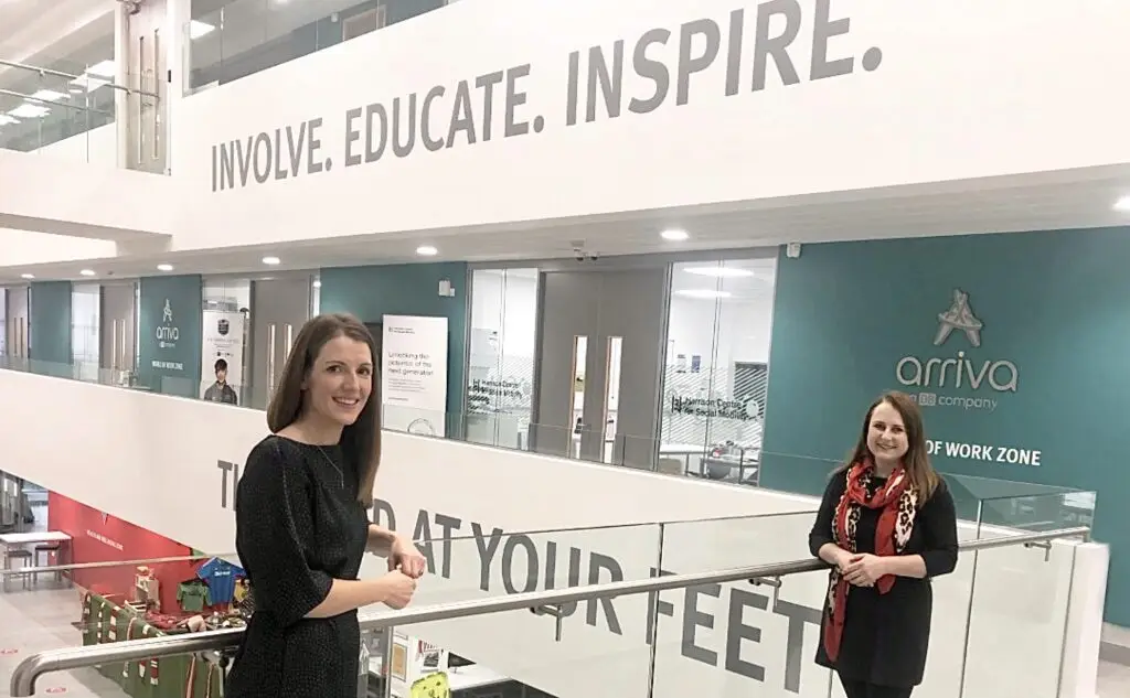 Bright photograph taken inside the Beacon of Light, with the words involve, educate and inspire visible on the walls. Two women stand against a glass partition smiling at the camera.