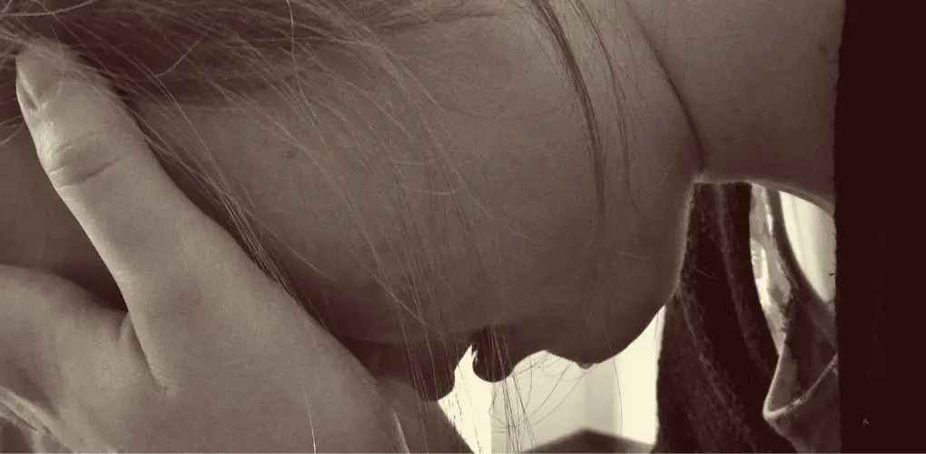 Extreme close up image of a woman's cheek, her eyes covered by hand hand and wisps of hair across her face. A tear is dripping from her chin. The photo has a sepia colour wash