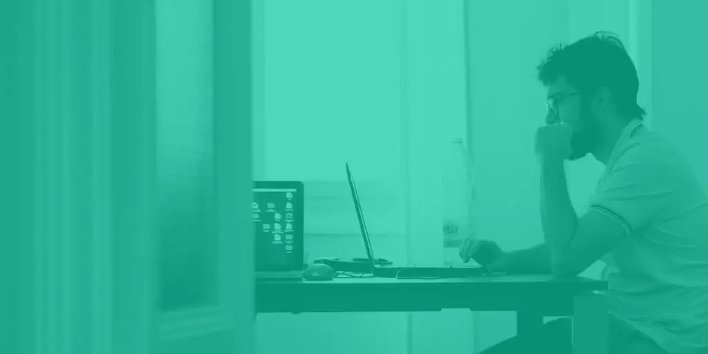Wide angle side shot of a man wearing glasses working on a laptop at a table. The full image has a heavy mint coloured hue over it.