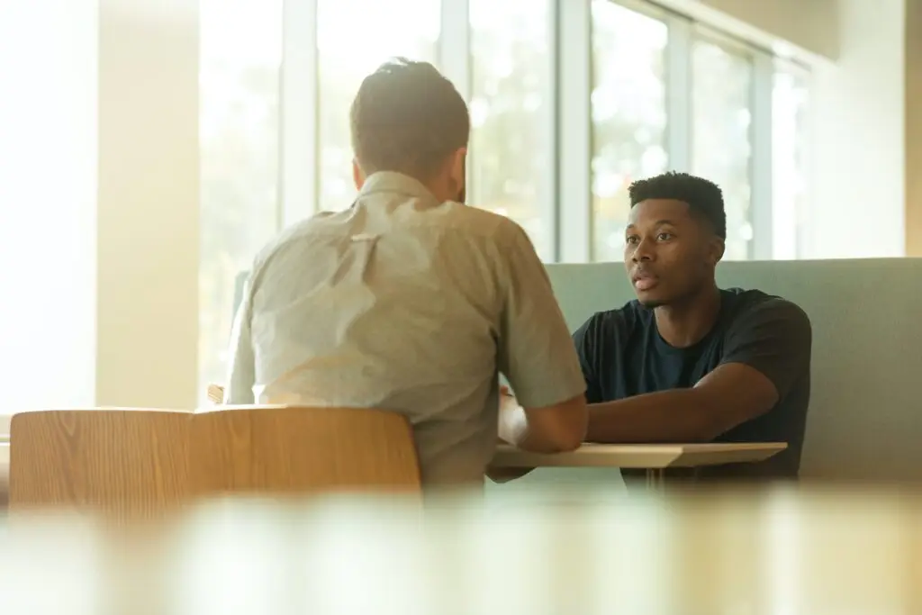 Close crop of two people talking while sitting at a desk facing each other. One has their back to the camera, they both sit close to a large window, the light from white casting a warm pale hue across the whole image.