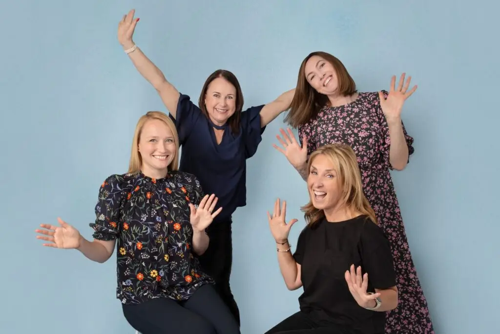 Photo of four women in playful poses and smiles, on pale blue background