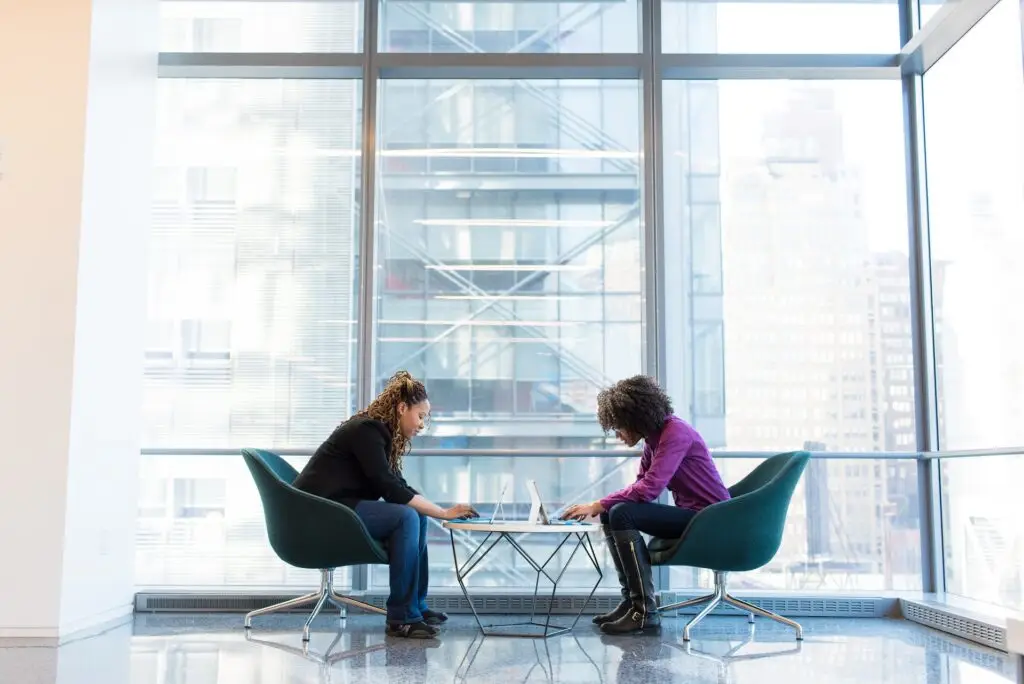 A side shot of two business women, sitting facing each other leaning over a small table, set again a large glass commercial wall.