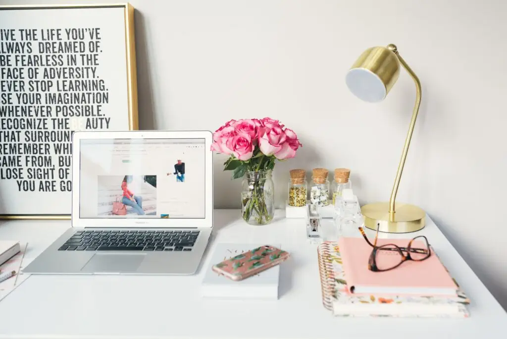 Close crop photo of a woman's desk, containing an large inspirational poster, an open macbook, pink roses, gold lamp, floral notebooks and smartphone, and a pair of glasses.