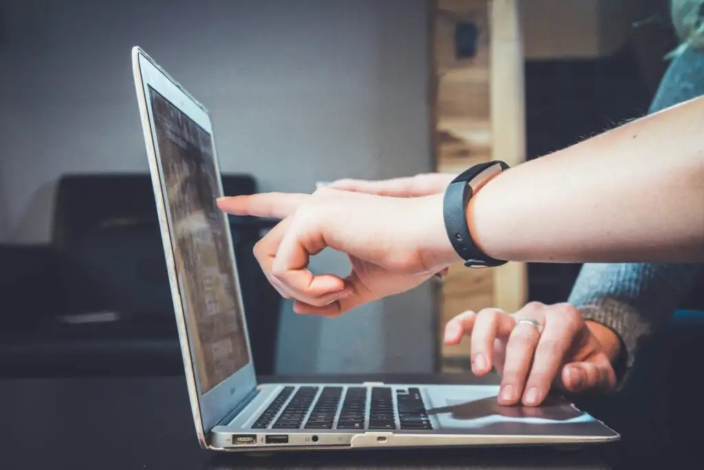 Close up side-shot photo of an open Macbook, one hand pointing at the screen with another resting two fingers on the mouse pad