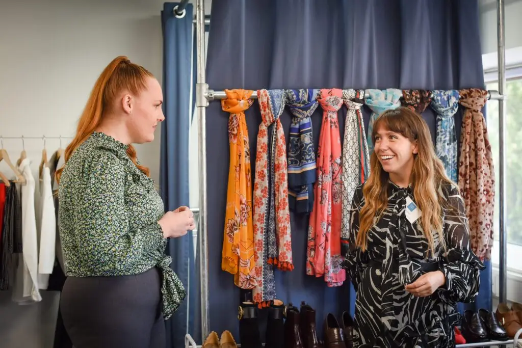 Photograph of two women in a room of clothing hanging up, casually talking to each other