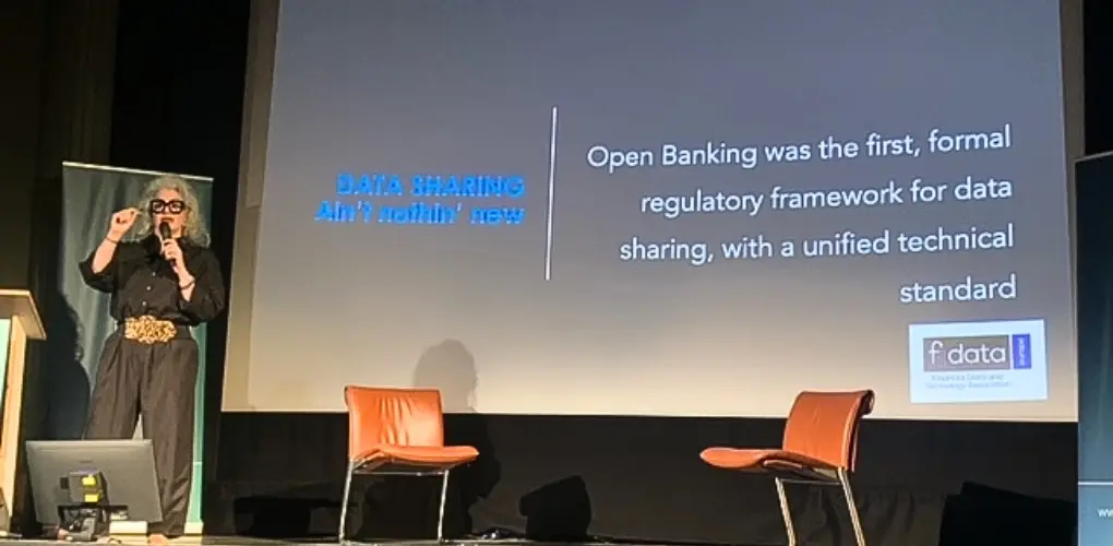 Photo of a woman with blond hair and black glasses on stage presenting about data sharing.