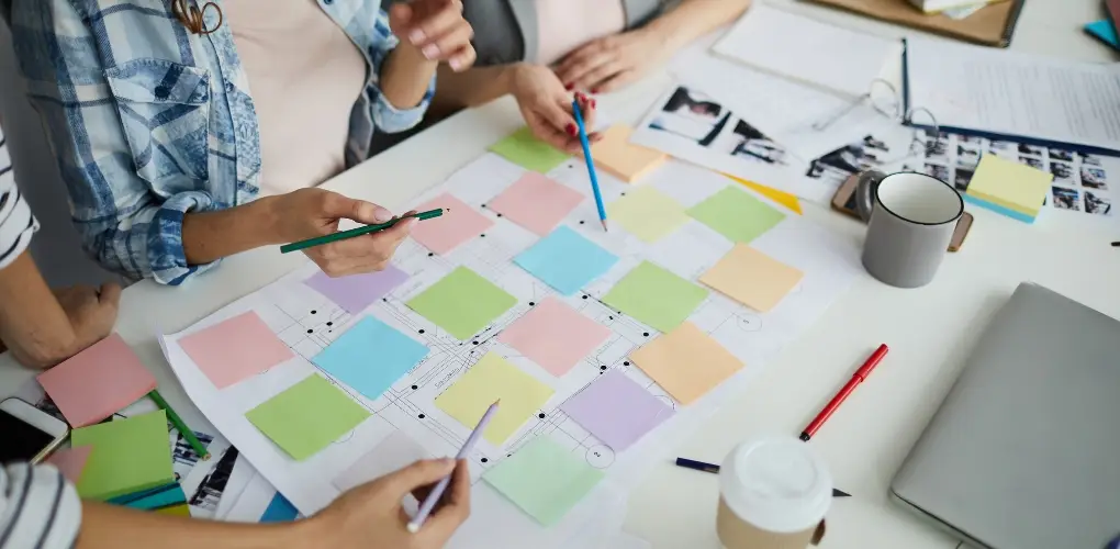 Close up photo of a desk covered in post it notes and drawings. Multiple woman surround the desk, although only their hands and pencils are visible.