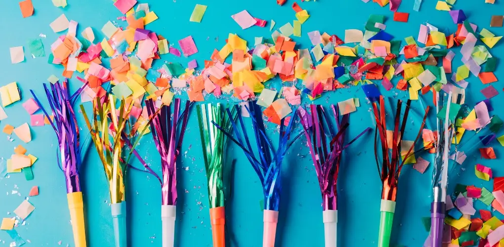 Photo of multiple tinsel whistles lined up on a blue background, surrounded by piles of multicoloured paper confetti.