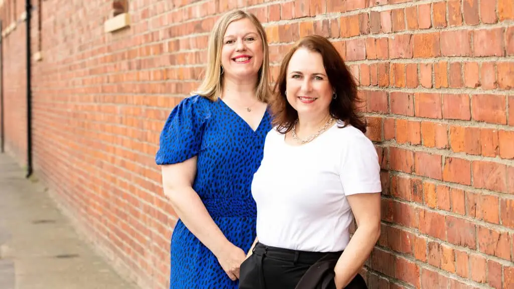 Close up of two MMC employees smiling at the camera at an outdoor location. They are stood overlapping, leaning against a red brick wall.
