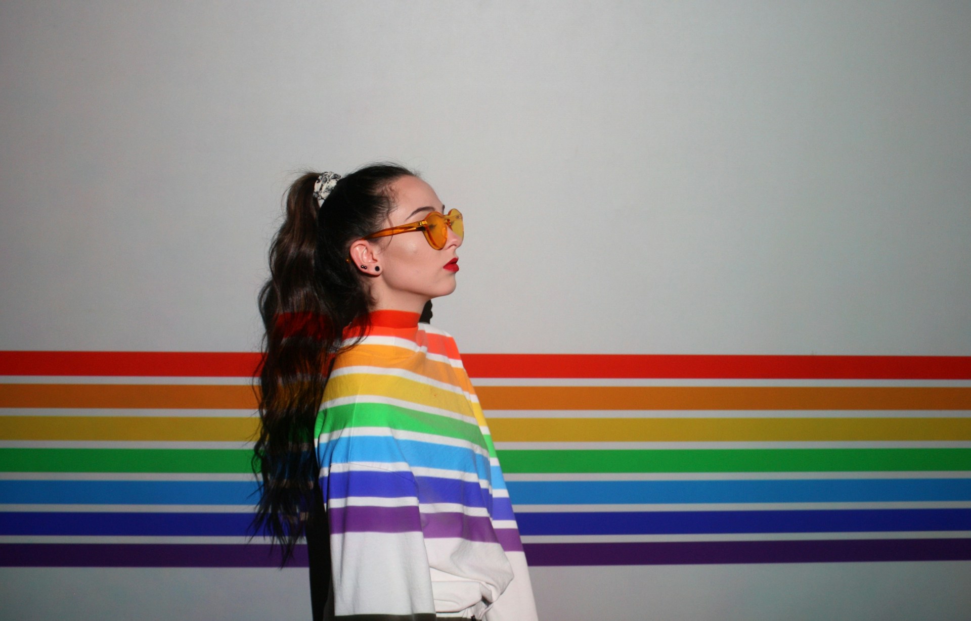 Photo of a woman wearing orange glasses standing sideways against a white wall with thin horizontal rainbow stripes. She wears a white long-sleeved top with matching rainbow stripes.