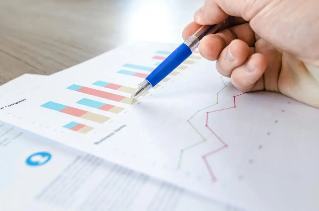 Close up photo of a hand holding a blue pen, pointing at multicoloured graphs on white paper.