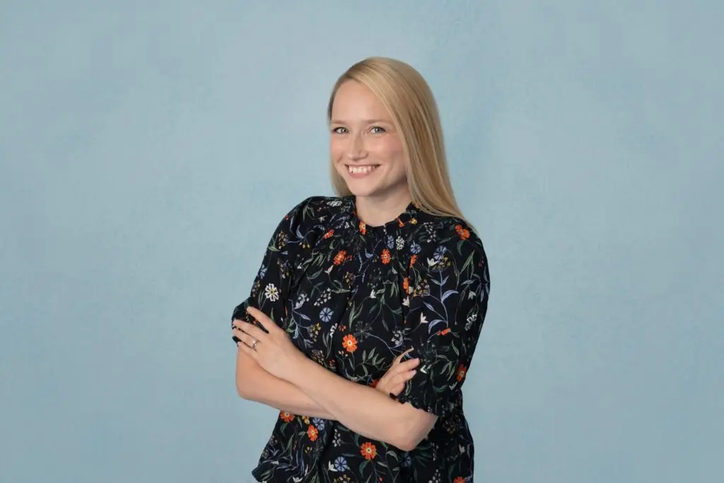 Photo of employee, Vicki Shouksmith, on a pale blue background, smiling at the camera with her arms closed.