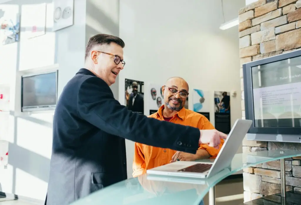 Photo of two men in a professional setting, smiling and pointing at information on computer screens.