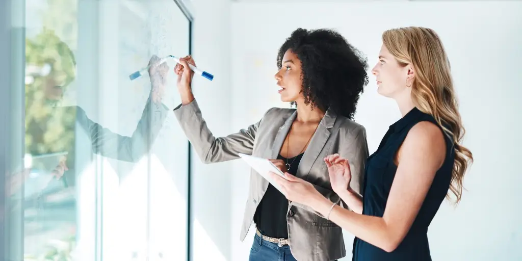Photo of two business women, shot from the waist up. They are shot from the side while writing on a clear whiteboard.