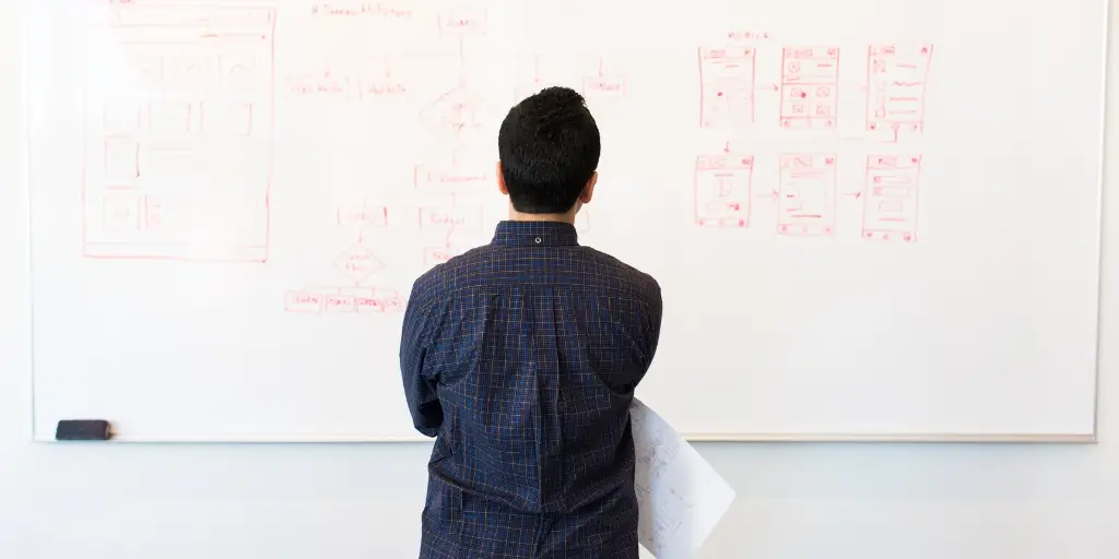 Photograph of a business man standing in front of a white board with vague illustrations in red pen